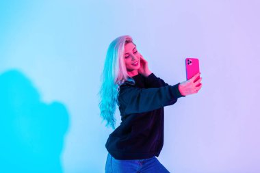 Siyah kapüşonlu ve kot pantolonlu mutlu güzel kız stüdyoda mavi pembe arka planda kendi fotoğrafını çekiyor. Güzel bir kadın akıllı telefondan selfie çeker.