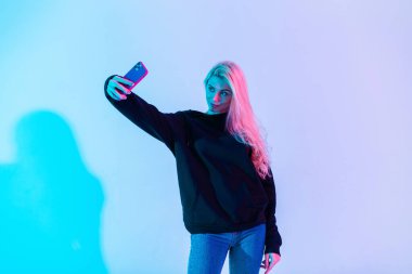 Moda kıyafetleri içinde güzel bir kadın kapüşonlu ve kotlu stüdyo telefonunda neon pembe ışık arka planına karşı selfie çekiyor.