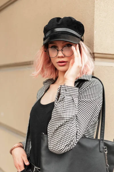 スタイリッシュなメガネと街のベージュの壁の近くにバッグスタンドとファッショナブルなプレイシャツの帽子を持つファッショナブルな美しい若い女性 — ストック写真