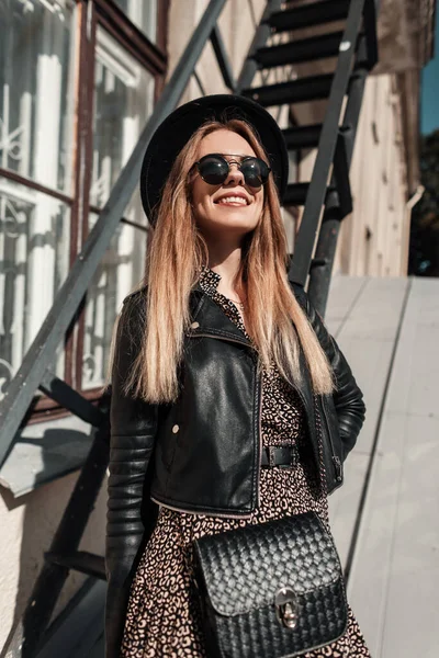 一个戴着帽子 戴着太阳镜 穿着黑色皮夹克 穿着古色古香的衣服 戴着时髦手提包 阳光照射在大街上的漂亮时髦女郎的艳照肖像 — 图库照片