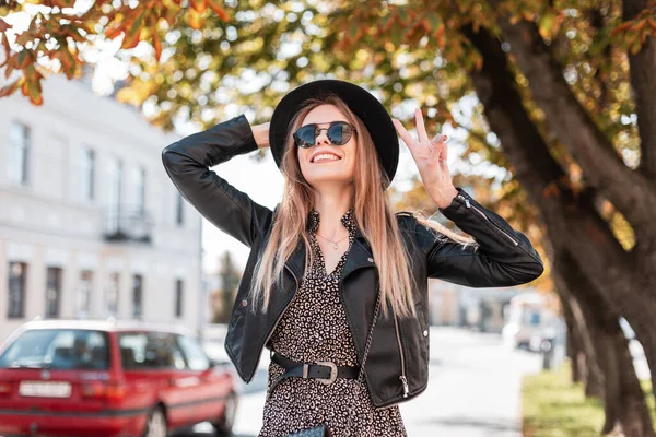 秋天的一天 一个穿着时尚皮夹克 穿着老式服装 戴着太阳镜 头戴一顶帽子的漂亮而滑稽的年轻女模特的时尚肖像在城市里走来走去 — 图库照片