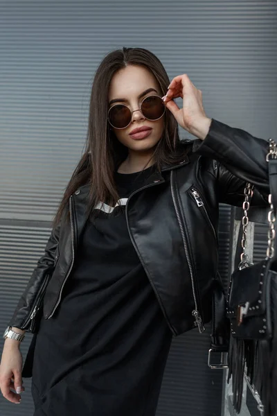 时尚漂亮的年轻女性模特穿着黑色皮夹克 戴着时尚的圆形太阳镜 穿着黑色时尚皮包的黑色连衣裙 站在城市的墙边 — 图库照片