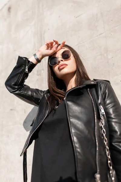 若い美しい女性モデルの女の子でファッショナブルなサングラスでスタイリッシュな黒革のジャケットは 太陽の光の下で晴れた日にコンクリート灰色のテクスチャの壁の近くに立つ — ストック写真