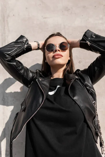 穿着时髦黑色皮夹克 汗衫的年轻貌美的女模特 戴着凉爽的太阳镜 站在阳光照射下的混凝土墙边 — 图库照片