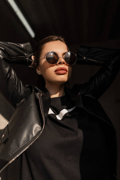 Модный творческий портрет красивой девушки с естественной красотой лица с большими сексуальными губами в винтажных круглых солнцезащитных очках с кожаной курткой позирует на улице с тенью и солнечным светом