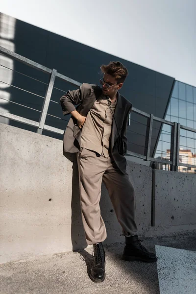 Cool young man model with vintage sunglasses in fashion gray blazer, shirt, pants and black boots stands on urban background at sunlight. Business casual style