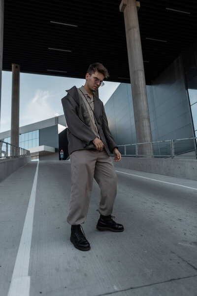 Стильная модель бизнесмена в старинных солнцезащитных очках в модном сером костюме с курткой, рубашкой, брюками и сапогами прогулки по городу