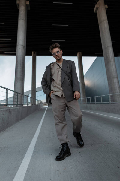 Стильный красивый хипстерский мужчина модель в модном наряде с блейзером, рубашкой, брюками и сапогами прогулки на фоне города.