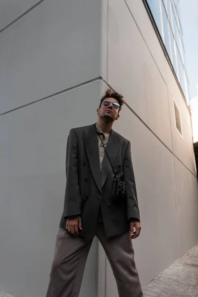 ファッショナブルな服でサングラスを持つハンサムなビジネスマンモデルのファッション肖像画は 市内の灰色の壁の近くに立っている — ストック写真