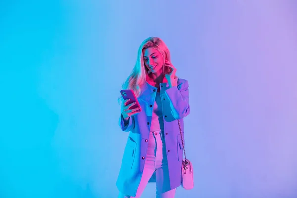 スタイリッシュなハンドバッグと明るいファッショナブルなビジネススーツの美しい若い幸せな女性は 電話を保持し 色のネオンピンクの光のスタジオでモバイルアプリケーションを使用しています — ストック写真