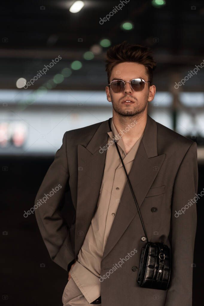 Portrait of stylish handsome business man with vintage sunglasses in fashion gray suit with handbag in parking