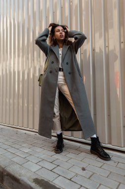 Şık, kıvırcık saçlı, şık ceketli, el çantalı ve ayakkabılı, sokakta metal bir duvarın yanında poz veren güzel bir kız.