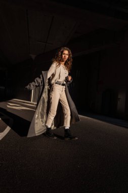 Örgü bluzu, pantolonu ve botlarıyla uzun paltolu güzel kıvırcık saçlı kız gün ışığında şehirde yürüyor.