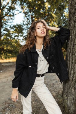 Şık, kıvırcık saçlı, şık siyah kot ceketli ve parkın kırsal kesimindeki bir ağacın yanında örülmüş kazaklı güzel bir kadın.