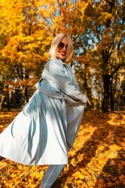 Güneş gözlüklü, sevimli bir gülüşü olan, hareketli mavi ceketli mutlu bir genç kız, sonbahar parkında sarı yapraklarla yürüyor. Güneşli bir sonbahar gününde kadınsı komik duygular.