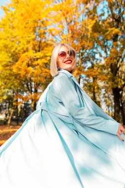 Kırmızı dudaklı, mavi elbiseli, güneş gözlüklü, mutlu, güzel bir kadın. Parkta parlak sarı sonbahar yapraklarıyla yürüyor.