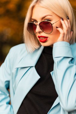 Güneş gözlüklü, kırmızı dudaklı güzel bir kadının parlak taze portresi gün batımında bir sonbahar parkında kameraya bakıyor.