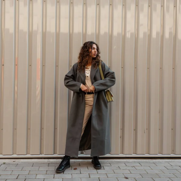 バッグ付きのヴィンテージグリーンのロングコートに巻き毛のファッションガールが金属の壁の近くに立っています 都会的なフェミニンなスタイルと美しさ — ストック写真