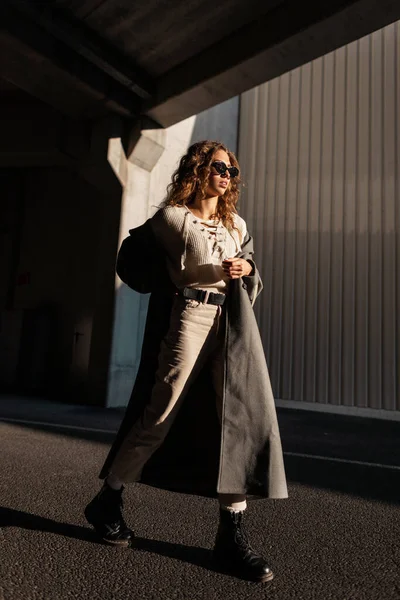 穿着时髦衣服的漂亮的卷曲女孩戴着太阳镜 穿着一件长长的外套在阳光下走在城市里 城市女性风格与美 — 图库照片