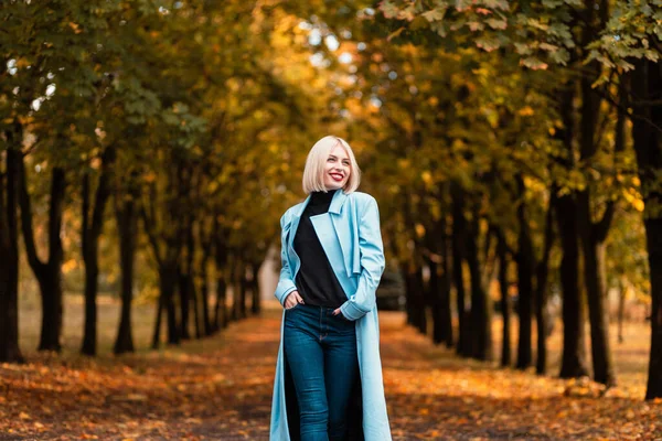 快乐美丽的女商人 穿着时髦的蓝色外套和牛仔裤 面带微笑 走在公园里 长满了秋天的树叶 — 图库照片