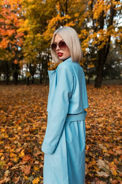 身穿时髦蓝色外套 戴着太阳镜 长着红唇的时尚而优雅而优雅的女人走在公园里 她的树叶是黄色的 — 图库照片