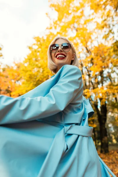 在秋天的公园里 身穿蓝色外套 戴着老式太阳镜 面带微笑的年轻而快乐的女人 走在一片金黄的秋天的公园里 — 图库照片