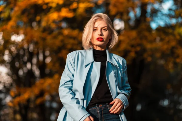 エレガントな若いですブロンド女性とともに赤い唇で青いコートの散歩で秋の公園でオレンジの葉で日没 — ストック写真