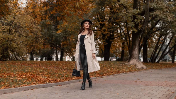 灰色の古典的なコート 黄色の葉を持つ秋の木々の近くの通りを歩くハンドバッグを備えたファッショナブルな服のエレガントな若い女の子 — ストック写真