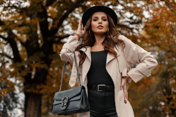 スタイリッシュな帽子と革の黒いハンドバッグを持つファッショナブルな服の中で巻き毛の美しい若いエレガントな女の子は 秋の葉を持つ木の背景を背景に通りを歩く — ストック写真