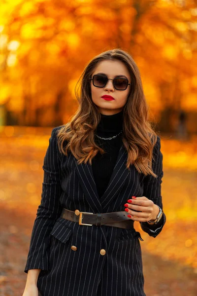 漂亮的年轻优雅的女商人 戴着老式太阳镜 穿着时髦的夹克 穿着毛衣 走在一个有橙色落叶的公园里 夕阳西下 女性秋风与美 — 图库照片