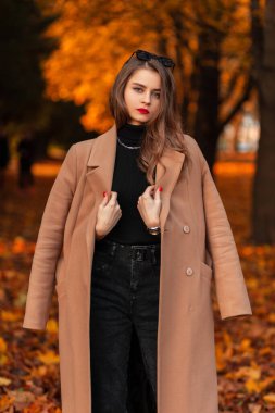 Güz parkında renkli turuncu yapraklarla şık bir bej ceket ve süveter giyen güzel, modaya uygun genç bir kadın yürüyor.