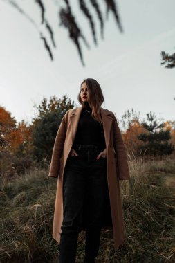 Genç, güzel bir kız model bej ceketli, çimlerin üzerinde siyah bir süveter ve pantolonu var.