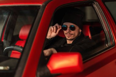 Havalı giyinmiş havalı bir adam eski arabasında oturur ve güneş gözlüğünü ayarlar.