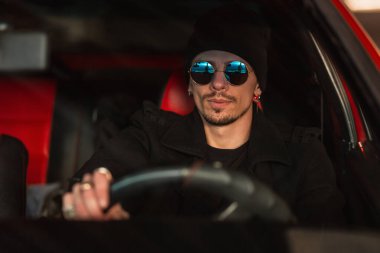 Güneş gözlüklü yakışıklı genç erkek sürücü kırmızı araba kullanıyor.