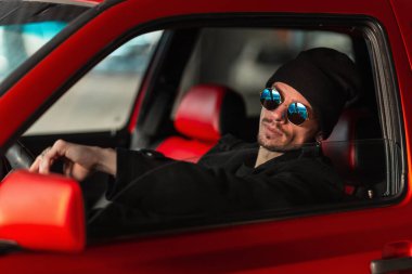 Güneş gözlüğü takan havalı genç bir erkek sürücü kırmızı bir araba kullanıyor. Adam direksiyonun başına geçiyor.