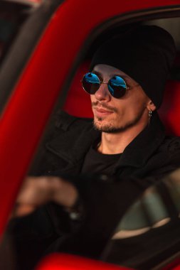 Şapkalı ve ceketli şık yakışıklı adam kırmızı eski bir arabanın direksiyonunda oturuyor.