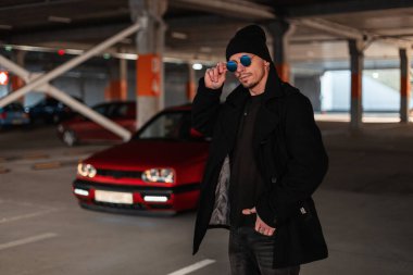 Siyah paltolu ve şapkalı yakışıklı genç adam park yerindeki kırmızı bir arabanın arka planına göre klasik güneş gözlüğünü ayarlıyor.