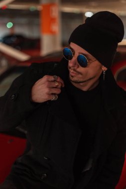 Şık, yakışıklı, güneş gözlüklü, siyah şık ceketli ve şapkalı genç bir adam.