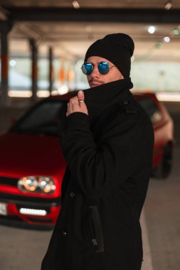 Moda güneş gözlüklü, şık bir adam ve park yerinde kırmızı arabanın yanında siyah, modaya uygun, şık bir ceket giyiyor.