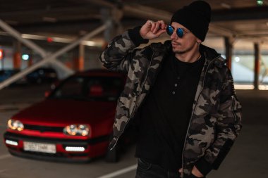 Havalı, genç, şık bir erkek modeli. Siyah şapkalı, şık bir asker ceketi ve kazağı var. Güneş gözlüklerini düzeltiyor ve şehre bakıyor.