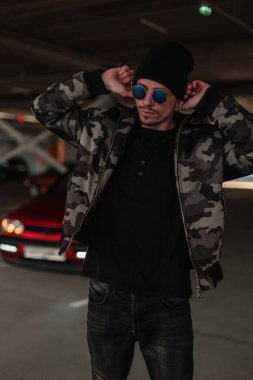 Moda askeri kışlık ceketli, güneş gözlüklü ve şehirde şapka takan yakışıklı bir adam modeli.