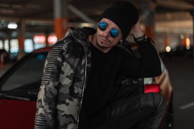 Şık güneş gözlüklü, siyah şapkalı, kışlık asker ceketli ve kazaklı yakışıklı bir gencin moda portresi. Kentsel erkek gündelik tarzı