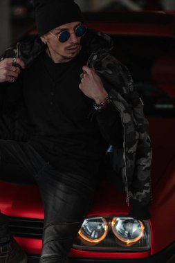 Moda askeri ceketli, şık güneş gözlüklü şık erkek modeli şehirde kırmızı bir arabanın yanında oturuyor.