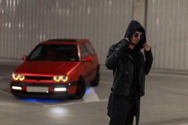 Havalı, yakışıklı, güneş gözlüklü, şık elbiseli, deri ceketli ve kapüşonlu bir adam bir gece parkında kırmızı akort yapan bir arabanın yanında poz veriyor.