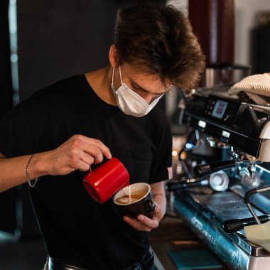 Siyah tişörtlü, tıbbi maskeli genç barista bir kafedeki kahve makinesinin yanında nefis kahve yapar. Coronavirus konsepti