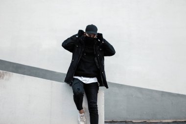 Şık kış ceketi ve kapüşonlu siyah şapkalı yakışıklı genç adam şehirde beyaz duvarın yanında koruyucu maske takıyor.