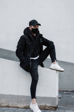 Siyah ceketli, şapkalı, kapüşonlu ve kot pantolonlu, koruyucu siyah maskeli şık genç adam duvarın yanında oturuyor.
