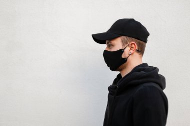 Koruyucu tıbbi maskeli, şık bir kapüşonlu siyah şapkalı, beyaz duvarın yanında yazı için boş yeri olan şık, yakışıklı bir genç. Gri arkaplanda erkek profili