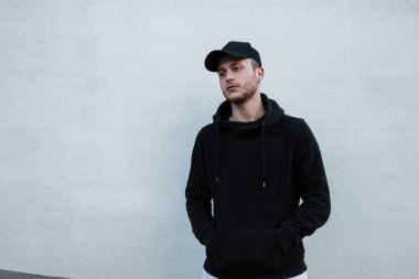 Havalı hippi siyah şapkalı ve şık siyah kapüşonlu adam sokakta beyaz duvarın yanında duruyor. Erkek kentsel tarzı