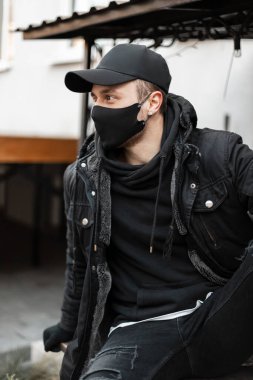 Siyah koruyucu maskeli, siyah ceketli, kapüşonlu ve şapkalı şık bir genç adam sokakta oturuyor. Kentsel erkek gelişigüzel tarzı ve koronavirüs konsepti